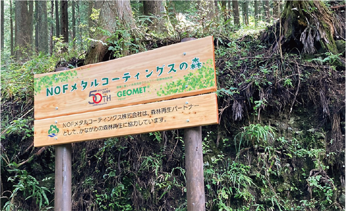 二本の丸太の柱に木の板がつけられて看板になっており、NOFメタルコーティングスの森とかかれています。