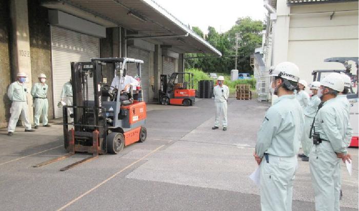 工場の倉庫の前の広い駐車スペースでフォークリフトの操作訓練をしています。回りに7，8名の作業員が訓練を見学しています。
