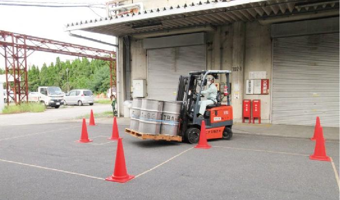 工場の倉庫の前の広い駐車スペースに赤いパイロンを並べて道を作って、フォークリフトの操作訓練をしています。