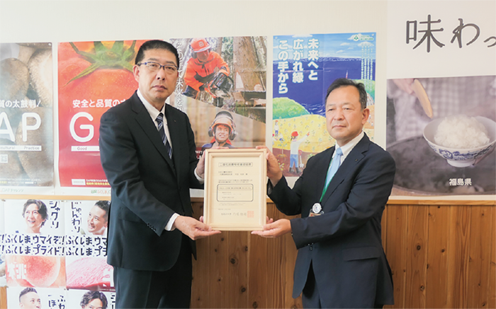ポスターが沢山貼られた壁を背に、表彰状を挟んで2人のスーツ姿の男性が立って記念撮影しています。