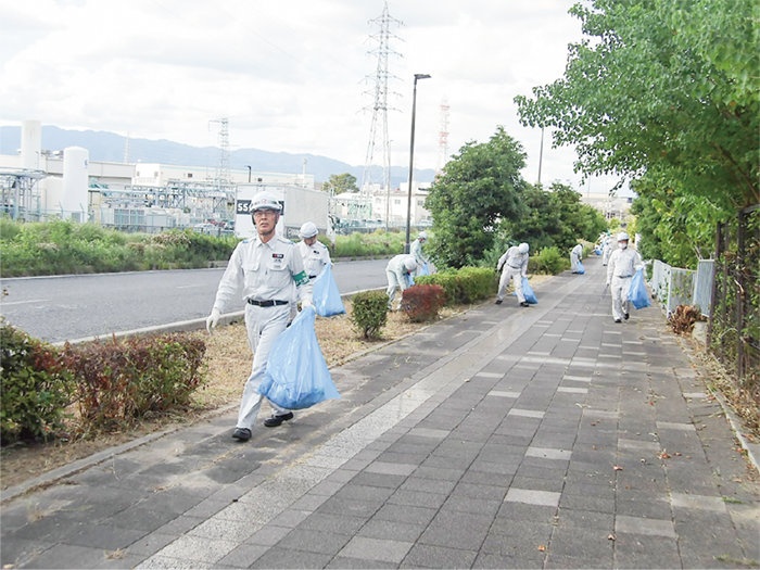 作業着を着た10数名の工場従業員が青いゴミ袋を持って、幹線道路脇の歩道の清掃作業をしています。