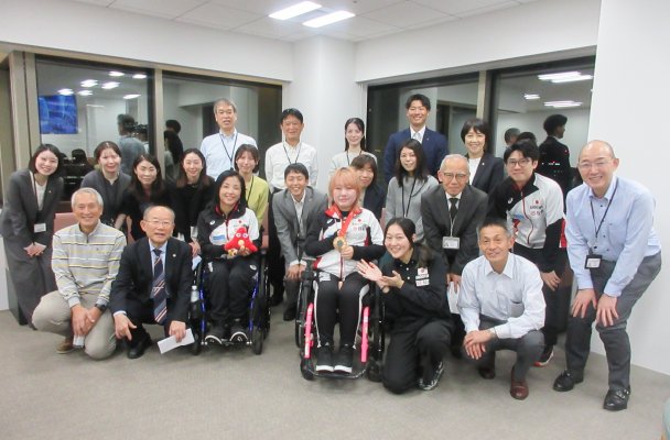 This is a group photo featuring Aki Taguchi, Head of the Japanese Delegation for the Paris 2024 Paralympic Games; Konaka Mizuta, bronze medalist in the air rifle prone event (SH2 classification for athletes with physical impairments); and employees of NOF Corporation.