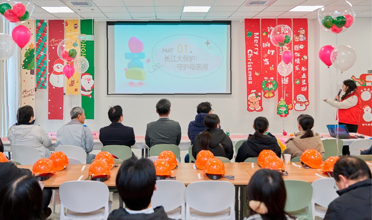 This is a photo of people gathered in a spacious meeting room decorated with balloons and Christmas ornaments, watching environmental education materials projected on a screen.