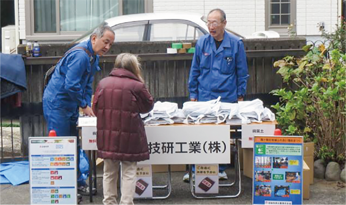 地域のお祭りの会場の一画に、ブルーシートを敷いて長テーブルを設置し、その上に無料配布用の自家製腐葉土を入れた袋が山積みにされていて、ジャンパーを着た男性2人が配布の準備をしています。