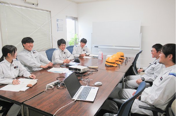 This is a photo of several young men in work clothes sitting side-by-side at a conference table, chatting in a relaxed atmosphere.