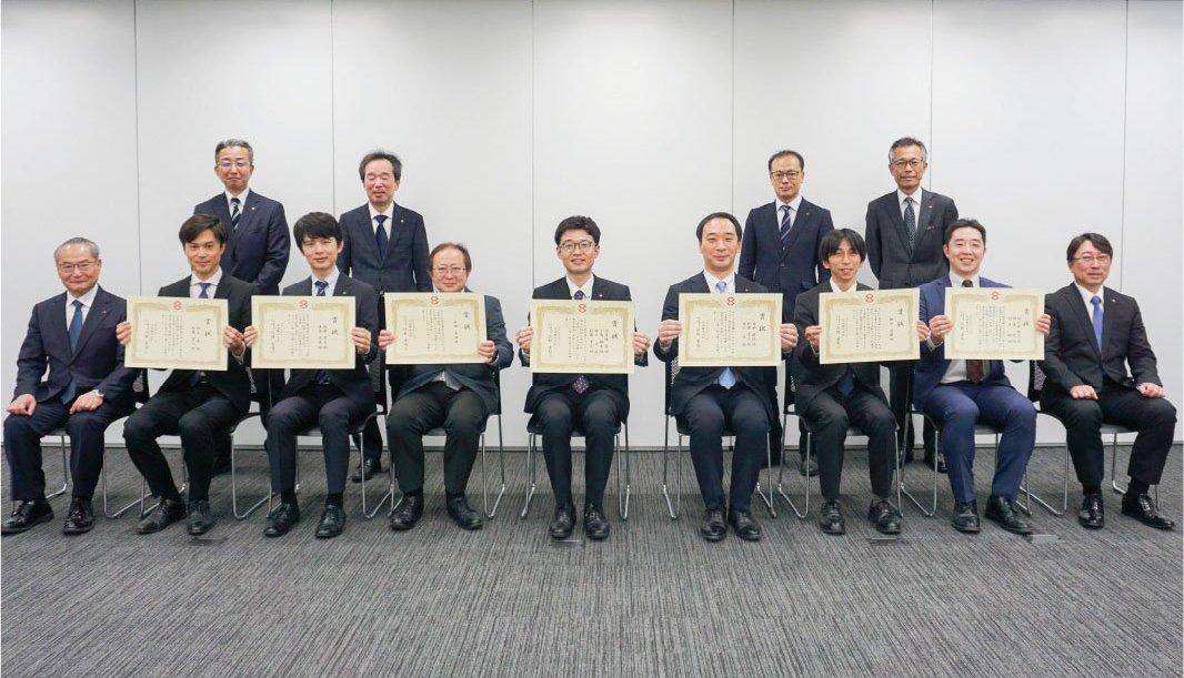 前後2列の集合写真です。褒賞された7人が賞状を持って前列に座り、その両サイドに会長と社長がそれぞれ座り、後ろに4人が等間隔に立っています。
