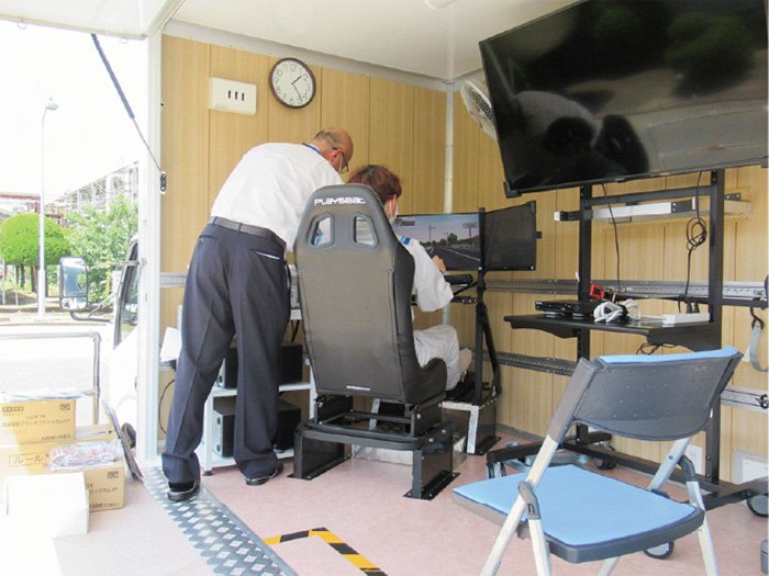 一人がバケットシートに座って自動車の運転シュミレーターで操作訓練をしています。別のもう一人が装置の操作をサポートしています。