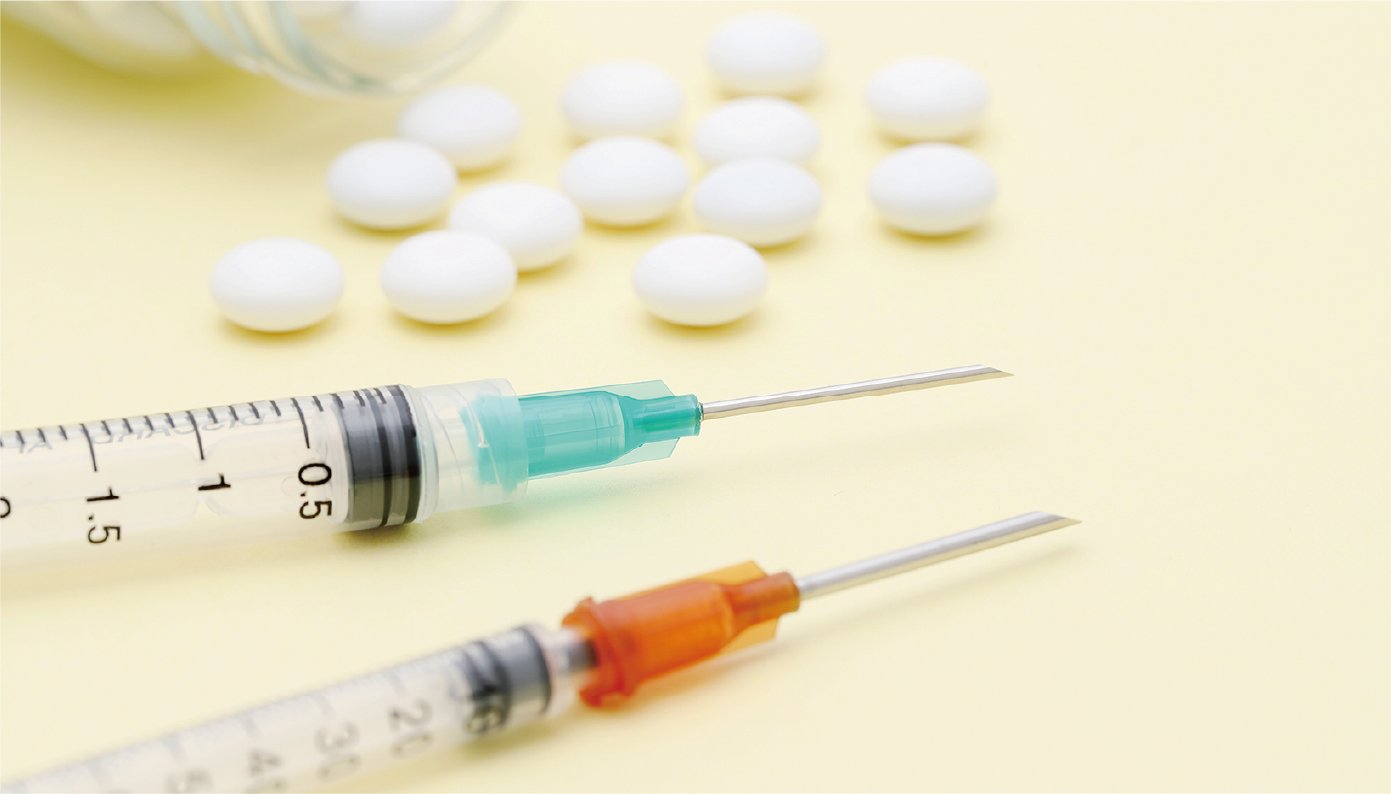 瓶からこぼれた白い錠剤が10数粒黄色いテーブルの上に広がっていて、黄緑色の注射器とオレンジ色の注射器が置かれています。