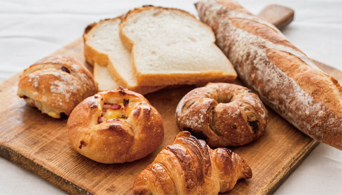 木製のトレーの上に、フランスパン1本、スライスされた食パン3枚、ベーグルのような丸形のパン3個、クロワッサン1個が並べられています。