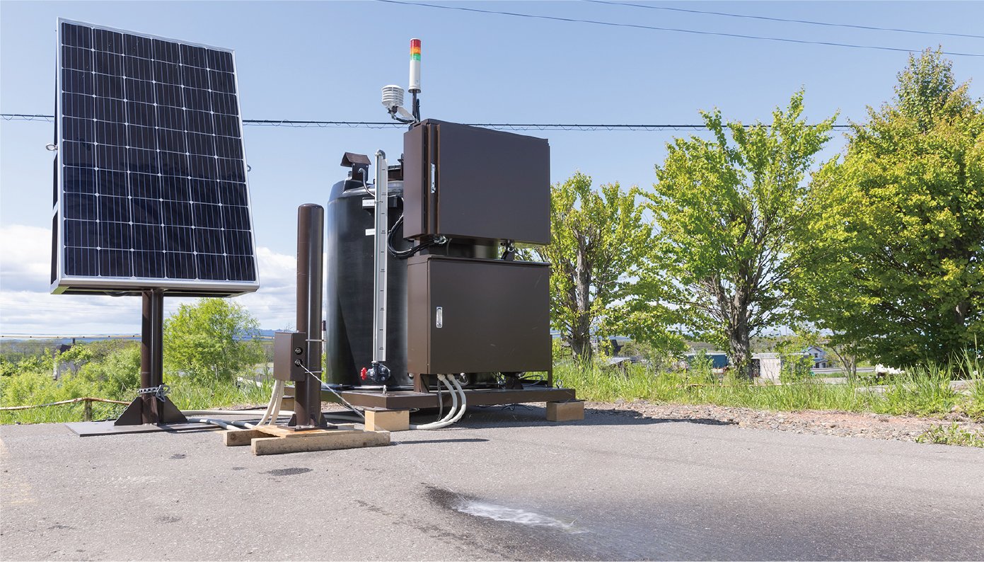 林の横を通る道路脇の開けた場所に機械が設置されています。その機械は、扉のついた茶色い箱が２つ手前についていて、中央にタンクがあって配管で繋がれています。すぐ隣に、動力となる太陽光発電パネルが設置されています。