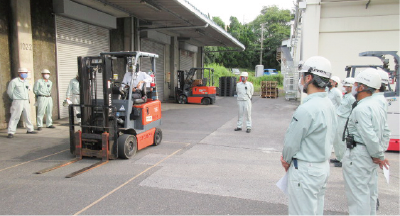 Forklift operation training is taking place in the spacious parking area in front of the factory warehouse. Seven or eight workers are gathered around, watching the training.