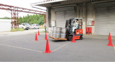 This shows a forklift training session at Nichiyu Logistics Co., Ltd. Within a course marked off by red traffic cones, a worker wearing a helmet and work clothes is operating a forklift to transport a pallet loaded with drums.