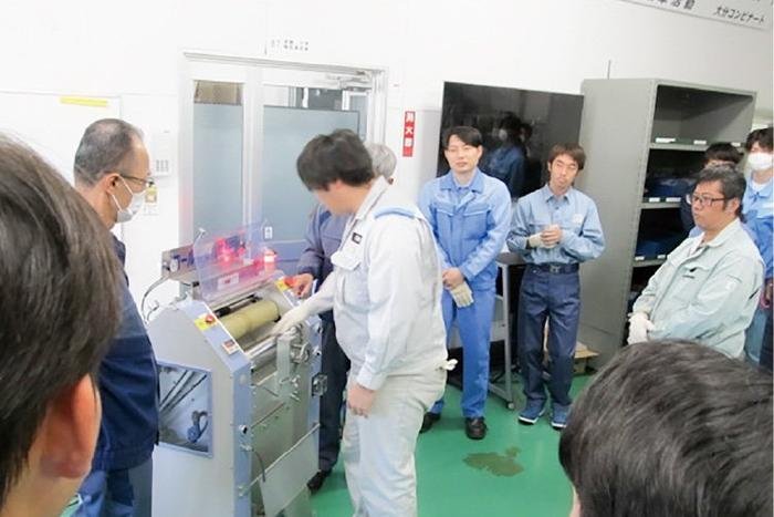 ロール型の危険体感装置に向かって、体験者の作業員が手に持った軍手がローラーに巻き取られてしまうのを体験しています。その回りで数名の人が様子を見学しています。