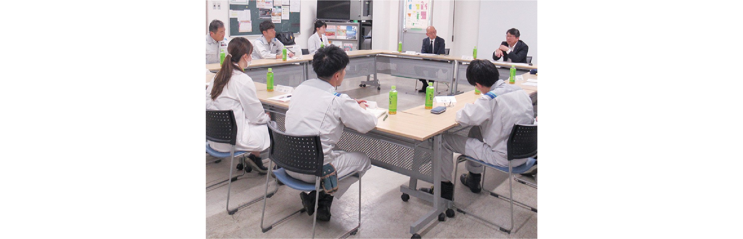 A town hall meeting is taking place around a table in a conference room. Takashi Sawamura, the President and CEO, is seated at the far end of the table, speaking with animated gestures, while employees dressed in work uniforms and lab coats listen attentively.