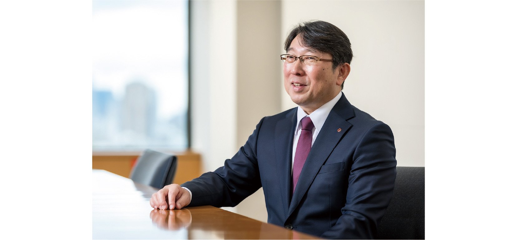 Koji Sawamura, the Representative Director, is speaking with a warm smile in a room featuring a large window and a cityscape in the background.