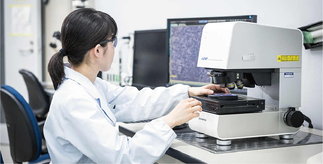 This shows research activities in the functional materials business. A female researcher is examining a specimen mounted on a microscope by viewing the image on a screen.