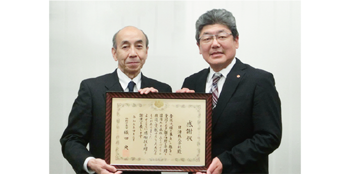 Two men in suits are taking a commemorative photo with a framed certificate of appreciation.
