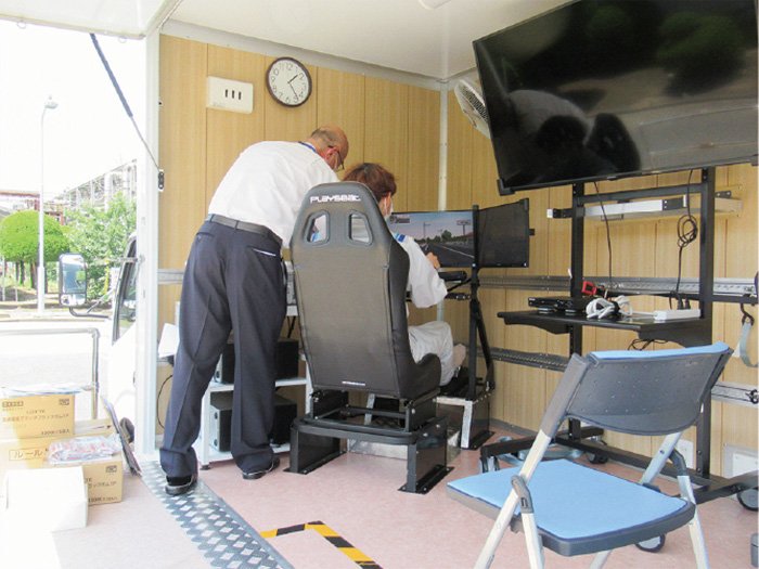 One person is sitting in a bucket seat and training to operate a car driving simulator. Another person is supporting the operation of the device.