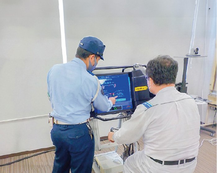 One person is sitting on a bicycle-style device and training to operate it using a bicycle simulator. Another person is supporting the operation of the device.