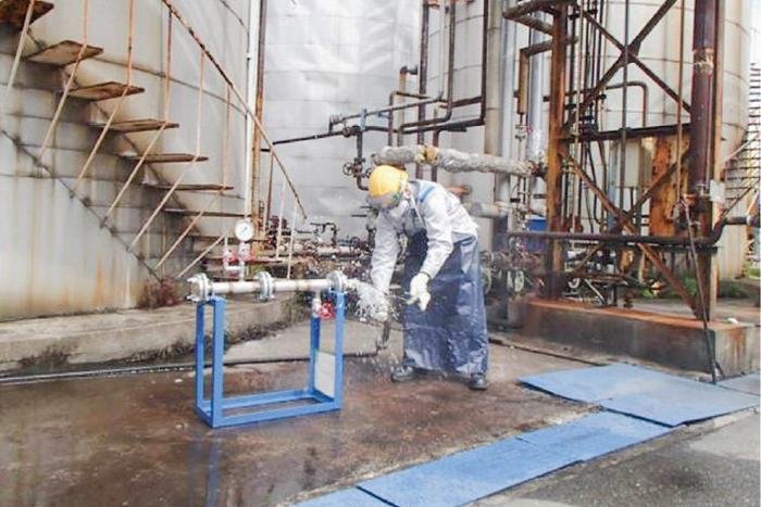 A worker wearing a helmet and protective gear is trying to disconnect a hose connected to a pipe, while facing a pipe-fixing-type hazard simulation training device.