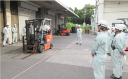 工場の倉庫の前の広い駐車スペースでフォークリフトの操作訓練をしています。回りに7，8名の作業員が訓練を見学しています。