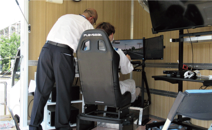 一人がバケットシートに座って自動車の運転シュミレーターで操作訓練をしています。別のもう一人が装置の操作をサポートしています。