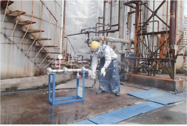 配管を固定した形の危険体感訓練装置に向かって、ヘルメットを被った1人の作業員が、防護具を付けて配管に繋がったホースを外そうとしています。