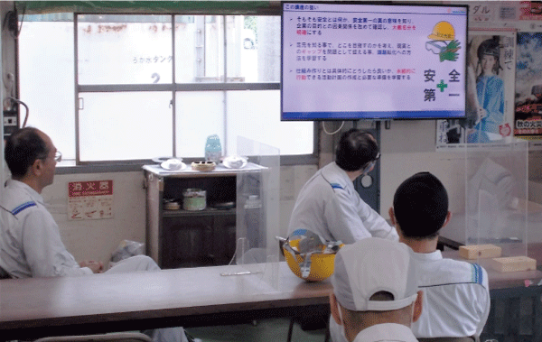 数人の作業服を着た人達がスクリーンに投影された安全教育資料を見て勉強会をしています。