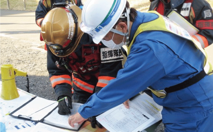 防災訓練で、屋外に設置された机に向かって、青い服に白いヘルメットで黄色のゼッケンを付けた報告役の人が、黒い服に黄色のヘルメットでオレンジの腕章と白い名札をつけた指揮役の人に、机の上の紙を指差しながら説明しています。