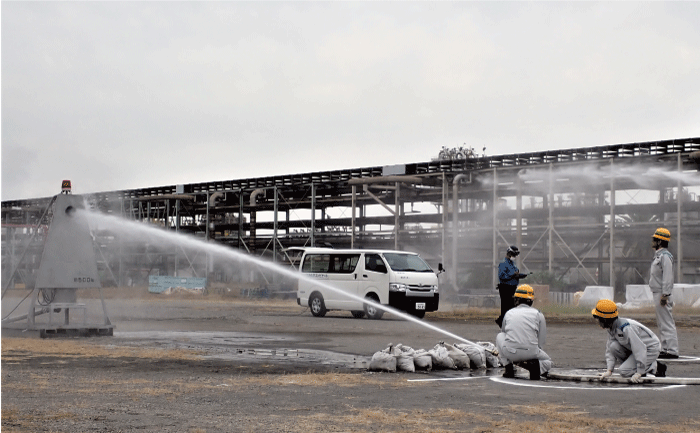 後ろに工場の配管ラックがあるとても広い空き地で、黄色のヘルメットで作業服を着た3名が、空き地の中ほどに、目印として立てられた旗をめがけて消火ホースで散水訓練をしています。