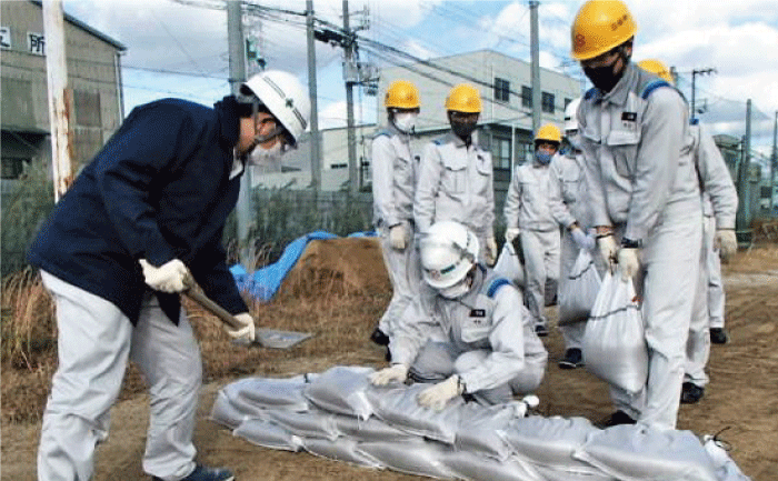 黄色のヘルメットをして作業服を着た5，6人が後ろで砂を詰めて土嚢を作っています。手前で白いヘルメットの作業員と黄色のヘルメットの作業員が土嚢を積んでいて、紺色のジャンパーを着て白いヘルメットを被った人がスコップで土嚢の壁に砂を寄せて壁を補強しています。