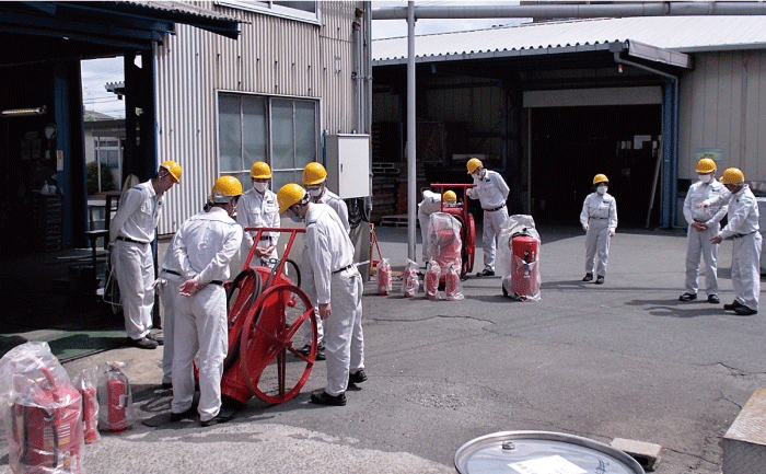 10数名の作業員が工場内の広場で消火器を並べて点検していて、左側に台車に乗せた大型消火器があり4人の作業員が取り囲んでいます。