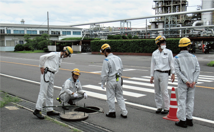 黄色のヘルメットを被った5人の作業員が、訓練で、工場の排水が流れ出す最終枡のマンホールを開けて中を確認しています。