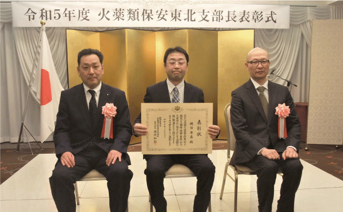 表彰式の会場で表彰されたスーツ姿の3人が、パイプ椅子に座って表彰状を正面に持って記念撮影しています。
