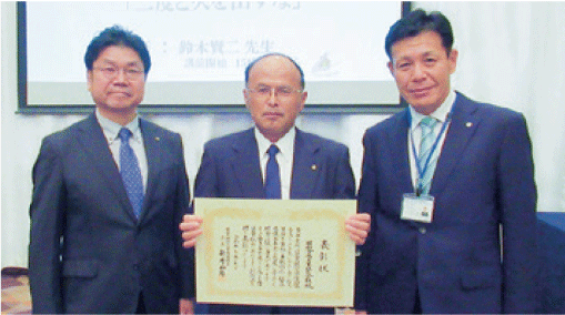 応接室の大きな額縁の絵を背景に、スーツ姿の3人が並んで立って、表彰状を正面に持って記念撮影しています。
