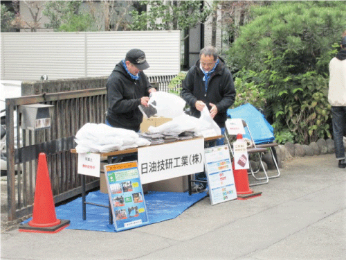 地域のお祭りの会場の一画に、ブルーシートを敷いて長テーブルを設置し、その上に無料配布用の自家製腐葉土を入れた袋が山積みにされていて、ジャンパーを着た男性2人が配布の準備をしています。