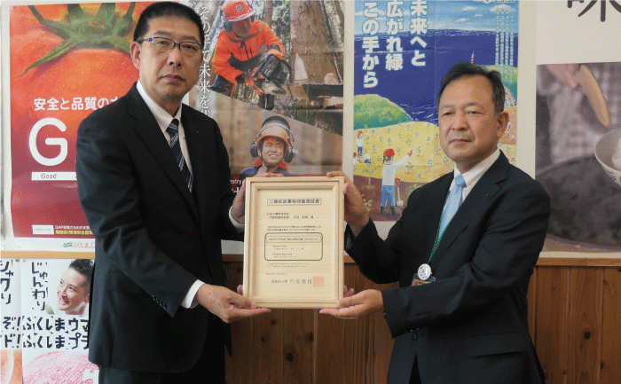 ポスターが沢山貼られた壁を背に、表彰状を挟んで2人のスーツ姿の男性が立って記念撮影しています。