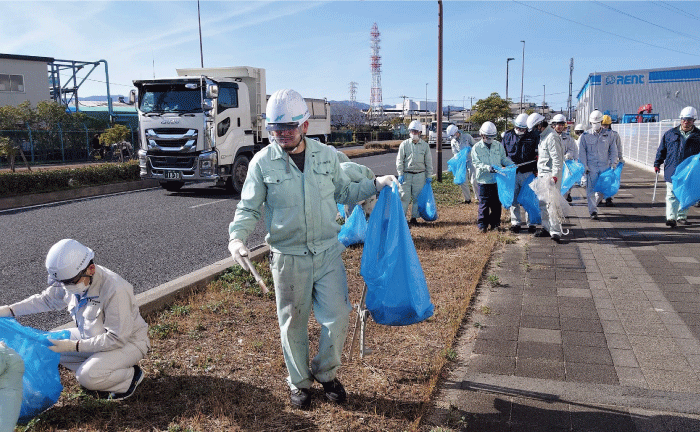 作業着を着た10数名の工場従業員が青いゴミ袋を持って、幹線道路脇の歩道の清掃作業をしています。