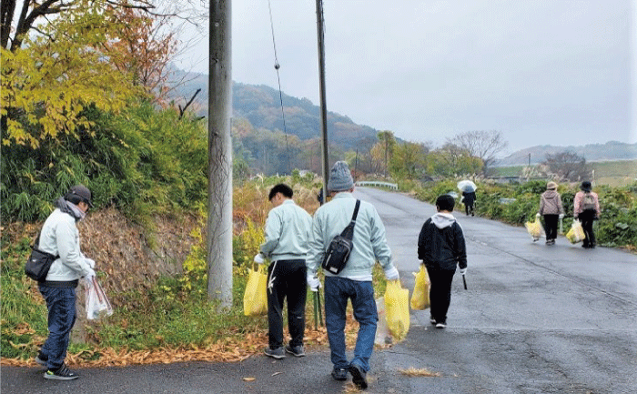 私服の数名の男女が山間の道路脇のゴミ拾いをしています。