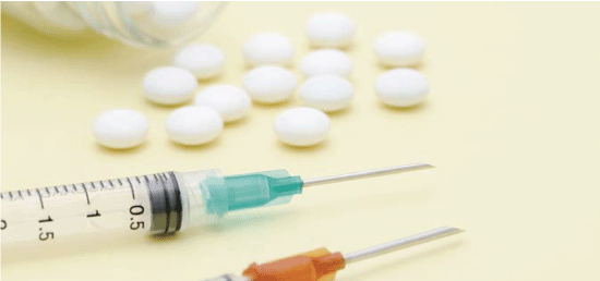 瓶からこぼれた白い錠剤が10数粒黄色いテーブルの上に広がっていて、黄緑色の注射器とオレンジ色の注射器が置かれています。